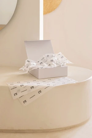 An open white presentation box lined with branded tissue paper resting on a curved beige pedestal in a minimalist interior setting.