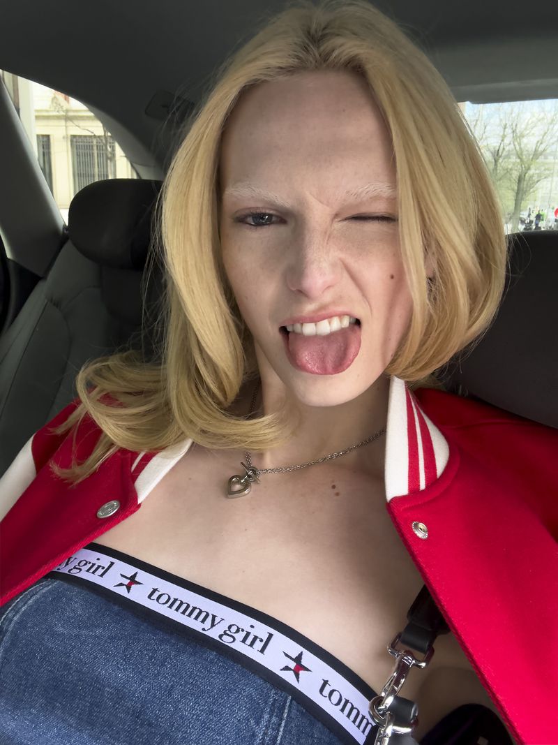 Close-up of a blonde woman winking with her tongue out, wearing a Tommy Girl crop top and red jacket, sitting in a car.