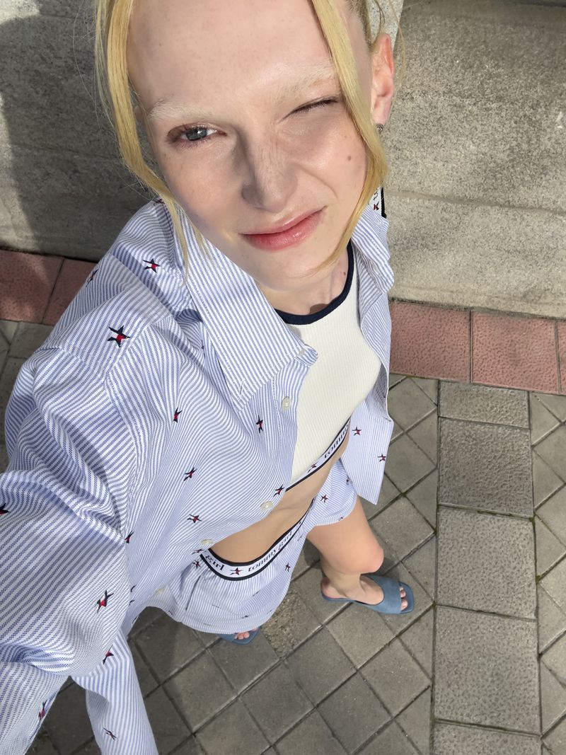 A young adult woman takes a selfie outdoors, wearing a blue and white striped shirt and shorts set with a matching white top, winking.