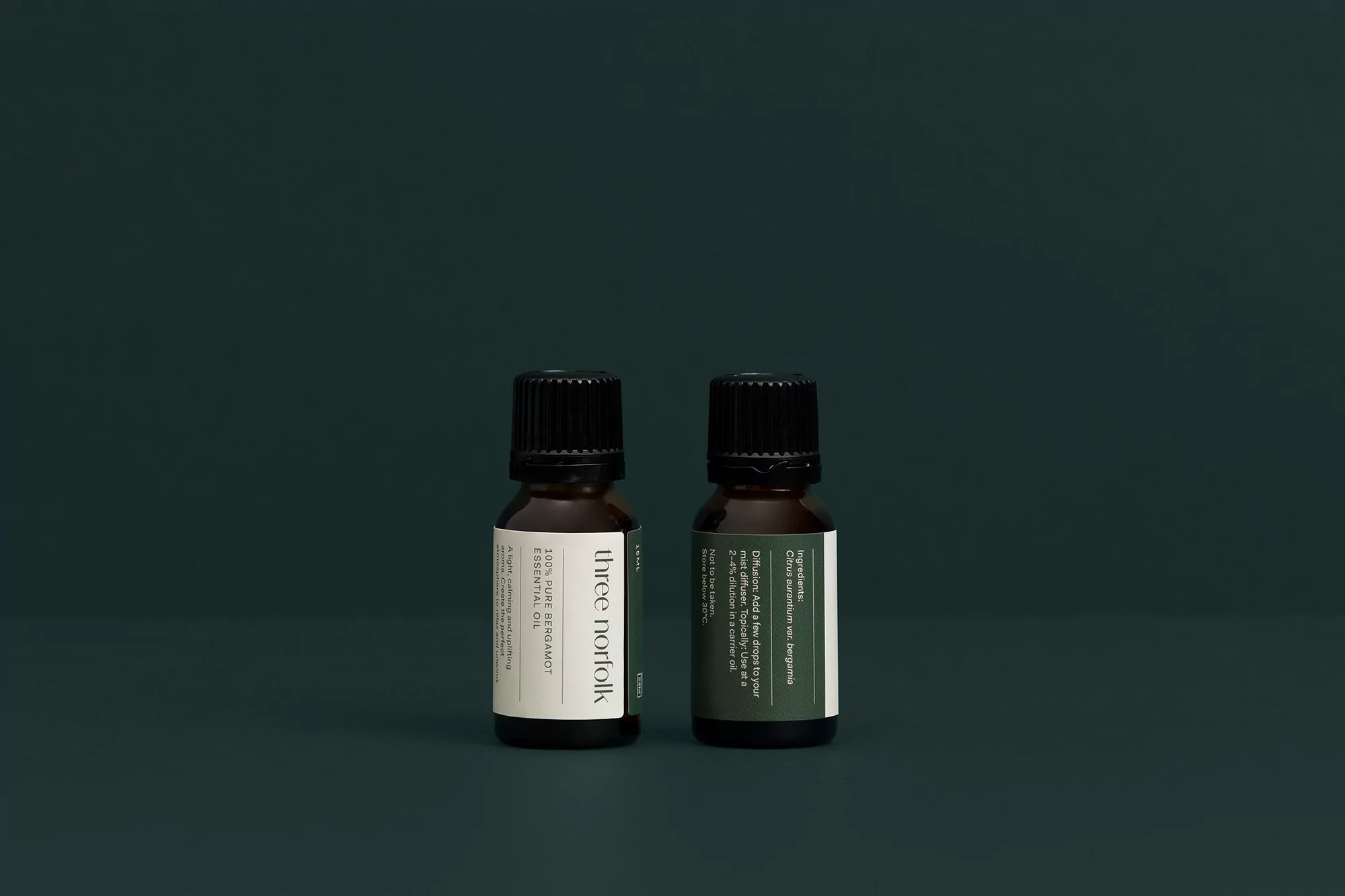Two small amber glass bottles of Three Norfolk bergamot essential oil stand against a deep teal background in a product shot.