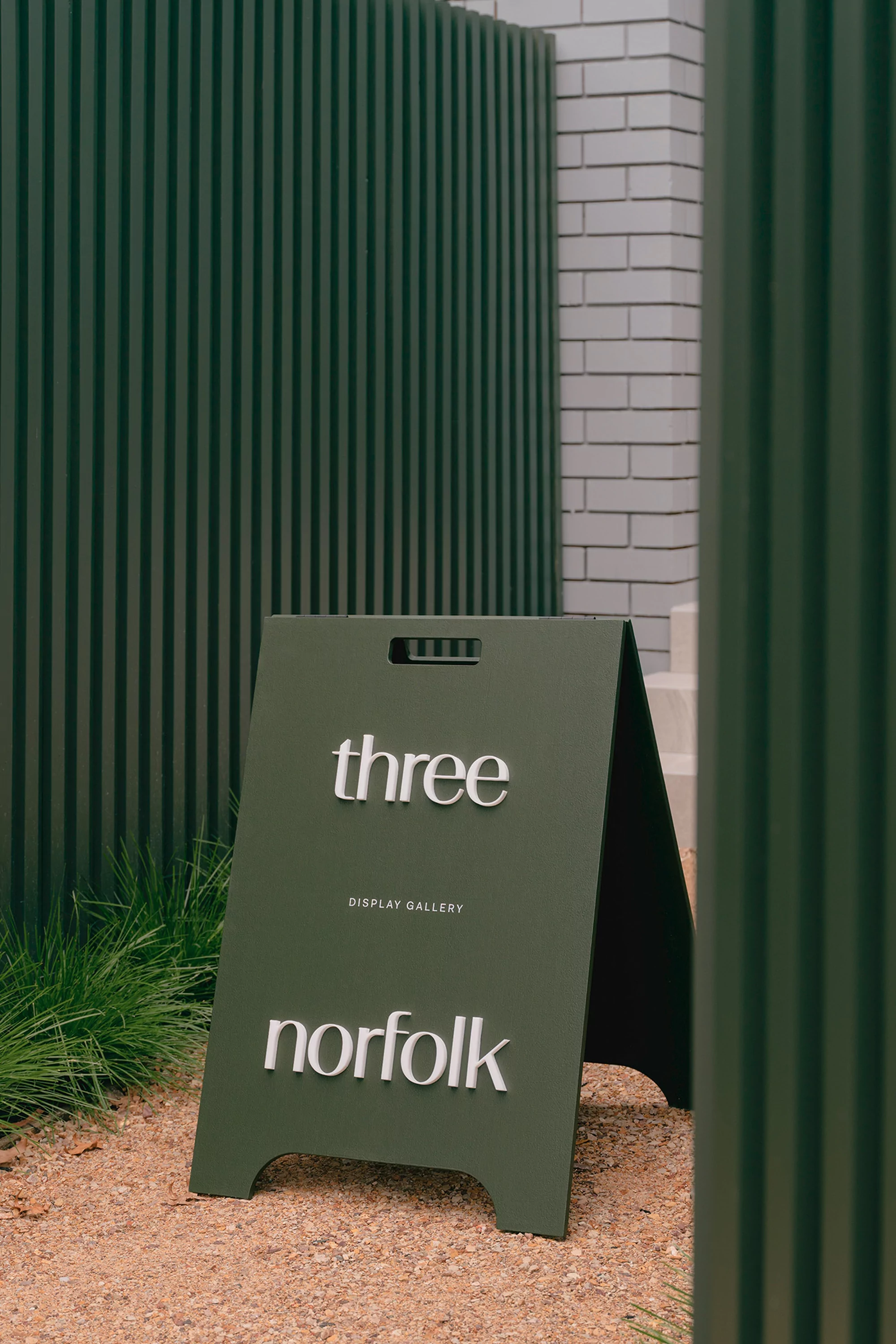 A-frame sign for Three Poplar Gallery in Norfolk, standing on gravel with green slat walls and a brick wall in the background.