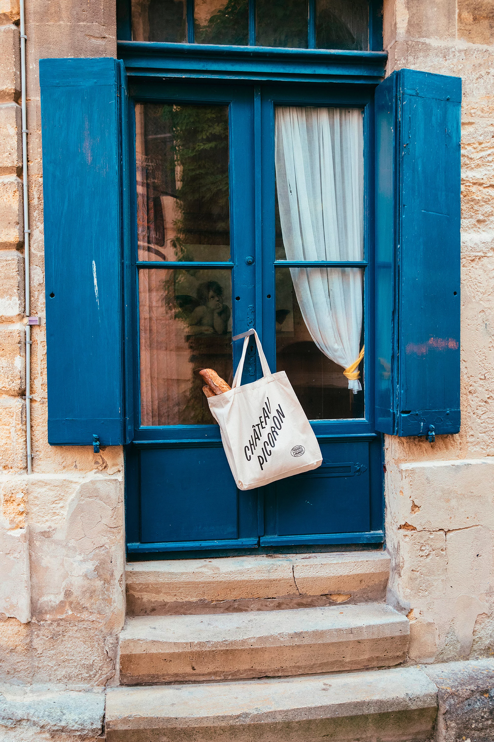 Blue shutters frame a French doorway with stone steps. A canvas tote bag with a baguette hangs on the door.