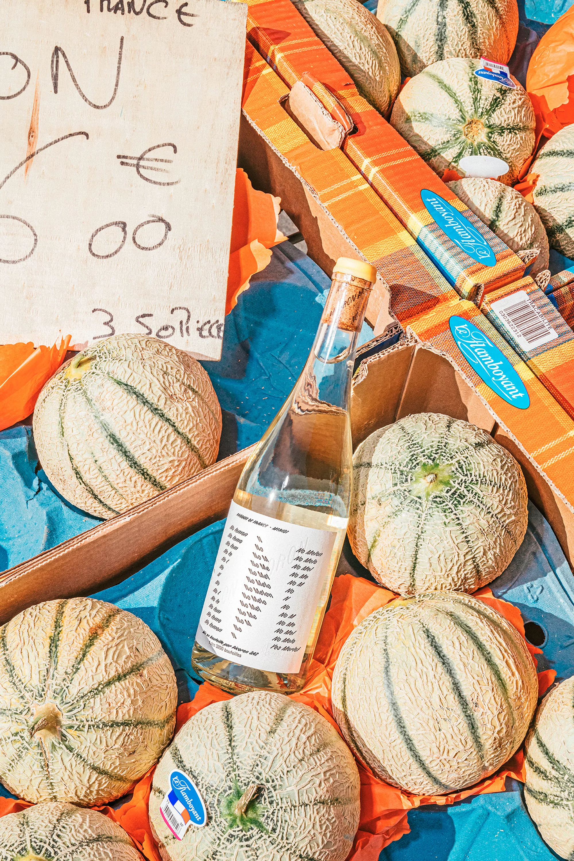 Melons are arranged in a box with a bottle and a sign at a food market, captured in a vibrant, eye-level outdoor shot.