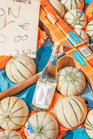 Melons are arranged in a box with a bottle and a sign at a food market, captured in a vibrant, eye-level outdoor shot.