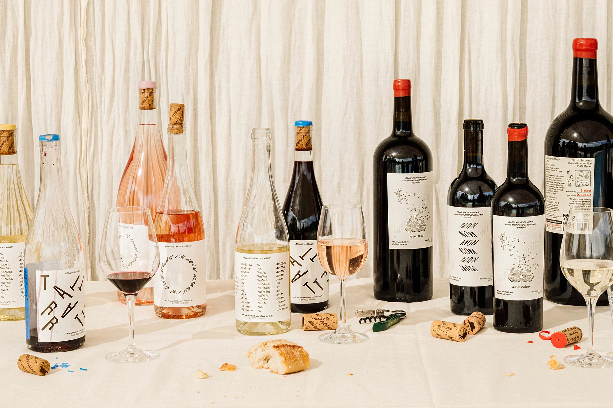 An array of wine bottles of varying colors and sizes are displayed on a white tablecloth in front of a neutral curtain backdrop.