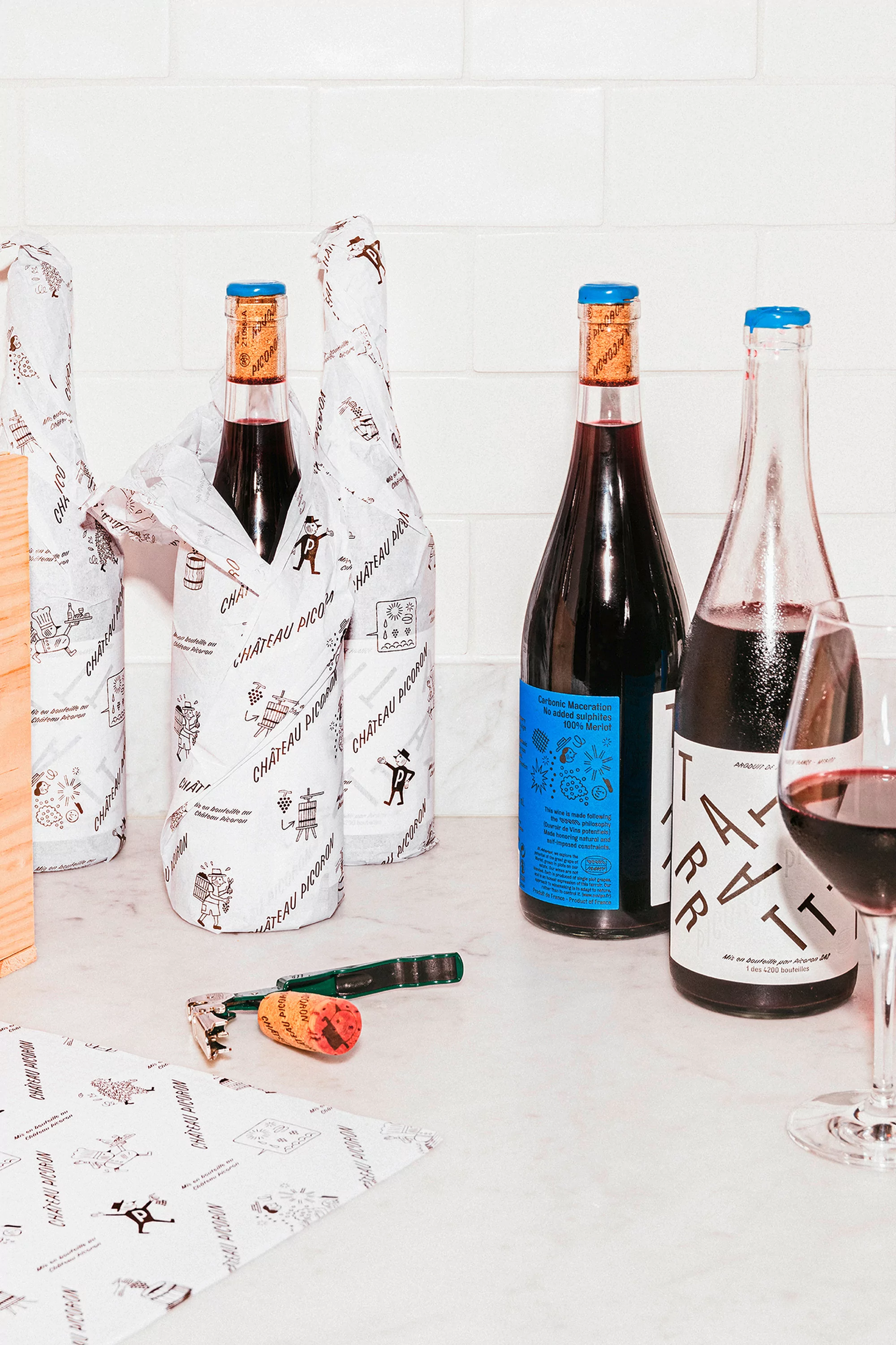 Bottles of red wine are on a white counter with a glass of wine, a corkscrew, and decorative paper against a white tile wall.