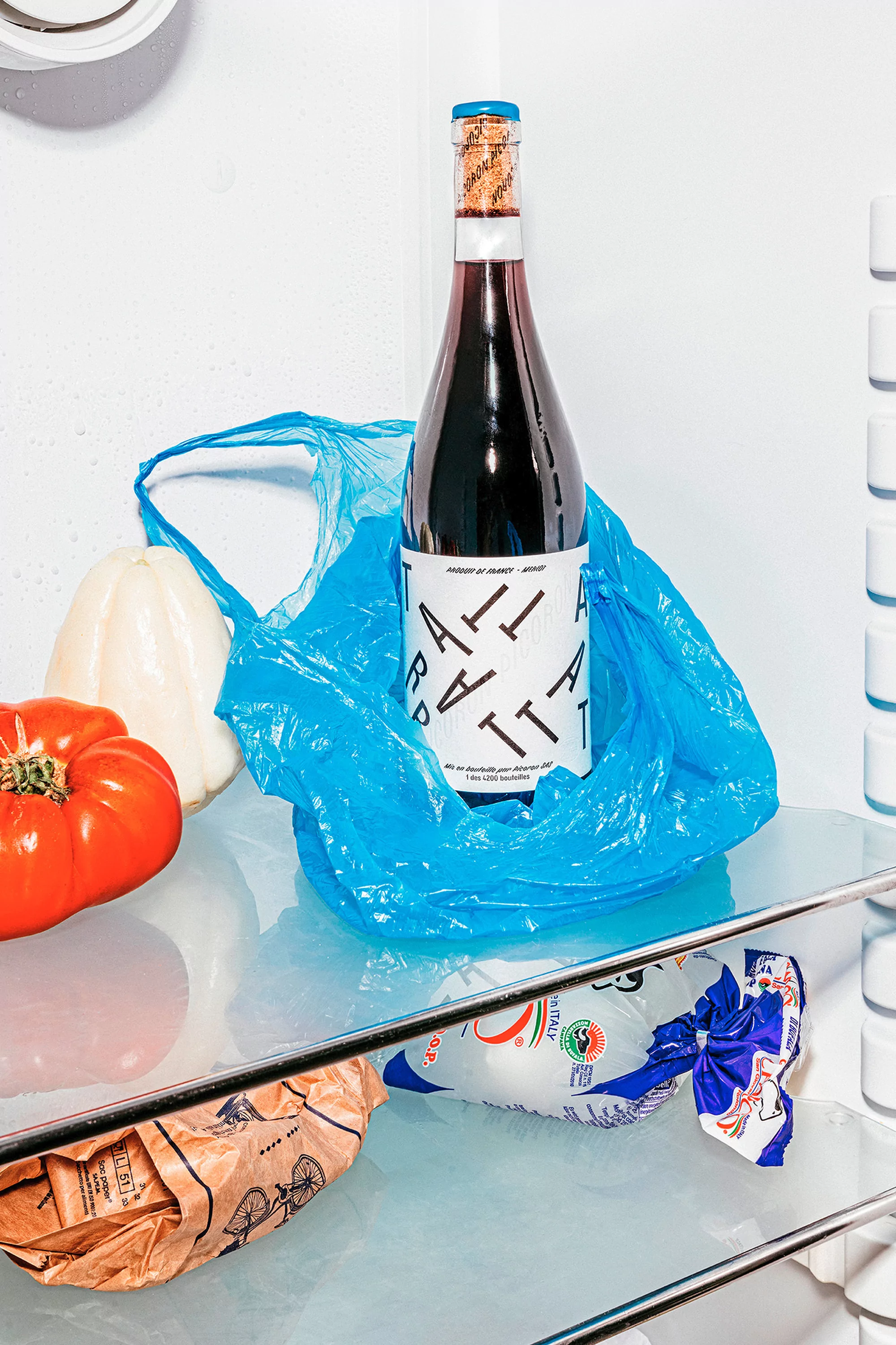 A bottle of red wine sits inside a blue plastic bag in a refrigerator, surrounded by tomatoes, peppers, and bread.
