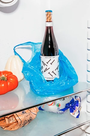 A bottle of red wine sits inside a blue plastic bag in a refrigerator, surrounded by tomatoes, peppers, and bread. 