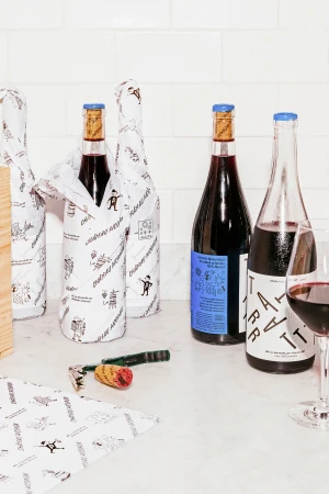 Wine bottles wrapped in illustrated paper and unwrapped next to a glass of red wine on a marble countertop.