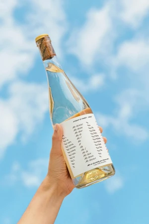 A hand holding a clear glass wine bottle with a white typographic label up against a bright blue sky with clouds.