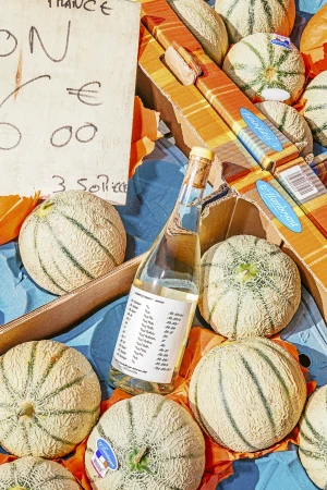 A bottle of white wine nestled among fresh cantaloupes in cardboard crates with a wooden market sign.