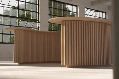 Two curved wooden slatted reception desks in an industrial showroom with large factory windows.
