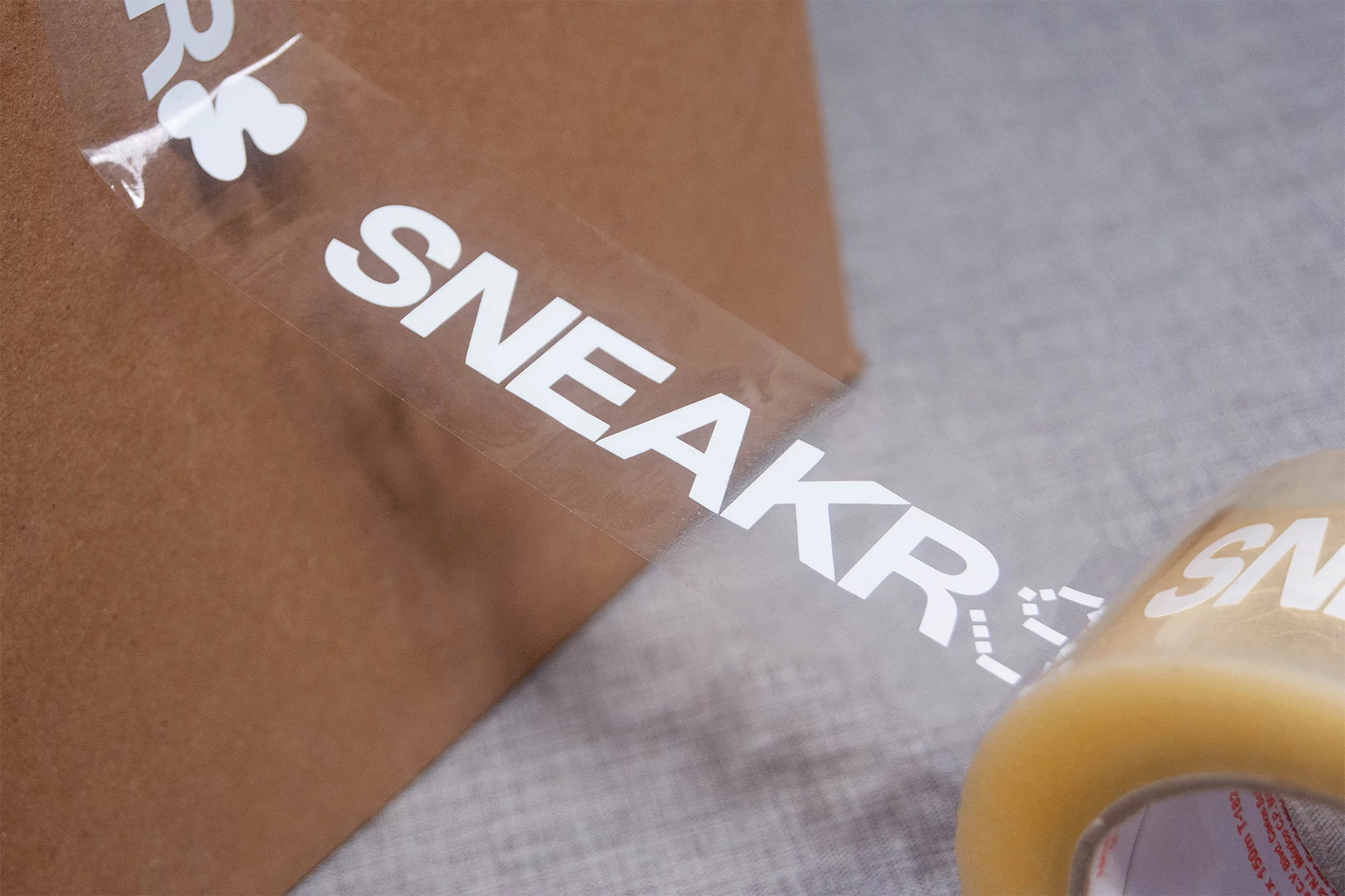 Close-up of transparent packaging tape with 'SNEAKR' branding on a brown cardboard box against a gray surface, product shot.
