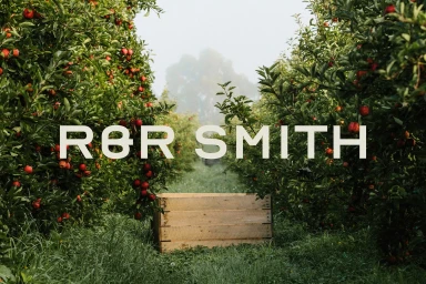 Wooden crate sits in the grassy path between rows of apple trees laden with ripe red fruit under an overcast sky.