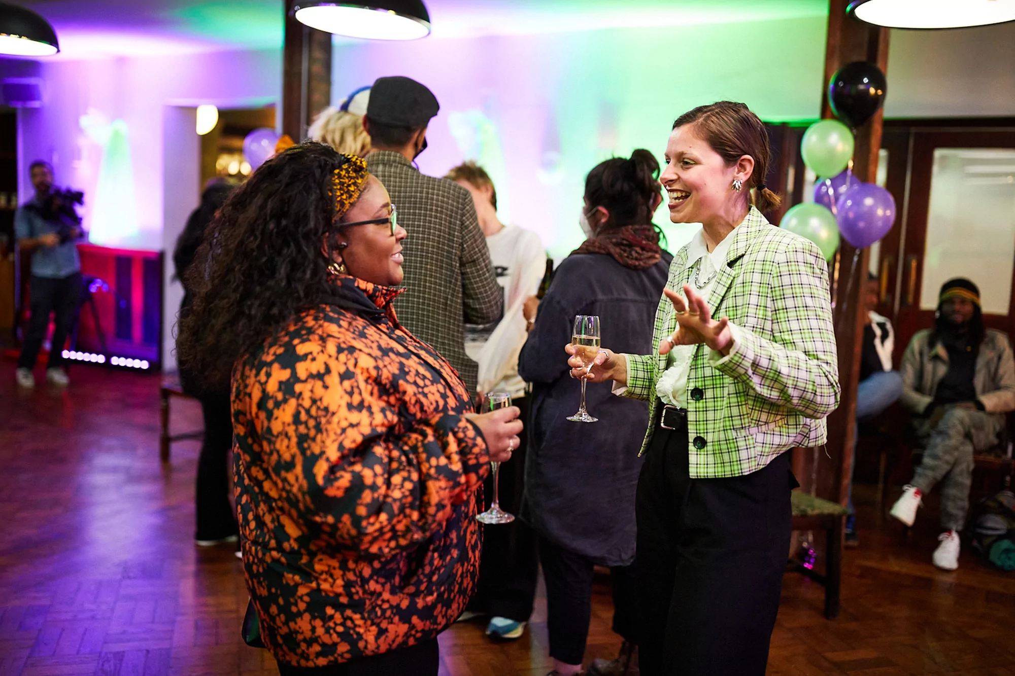Two adult women in a lively indoor gathering, holding champagne flutes, engaged in a conversation. Festive ambiance with balloons.