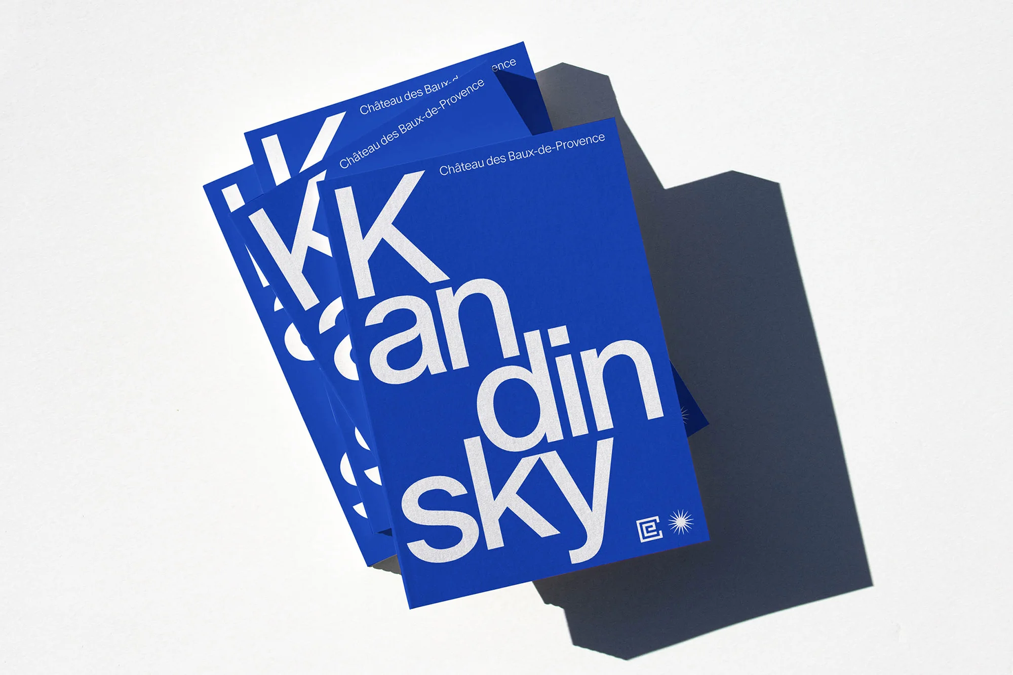 Three blue Kandinsky Château des Baux-de-Provence books are stacked on a white surface casting a dark shadow from the studio lighting.