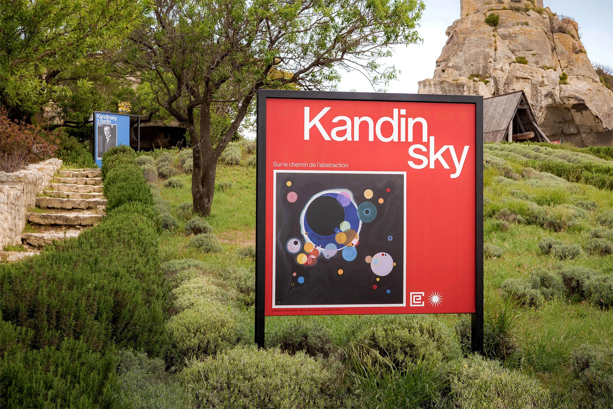 A large red Kandinsky Sky artwork display is outside, showing a colorful abstract circle painting, stone steps, and greenery on a sunny day.