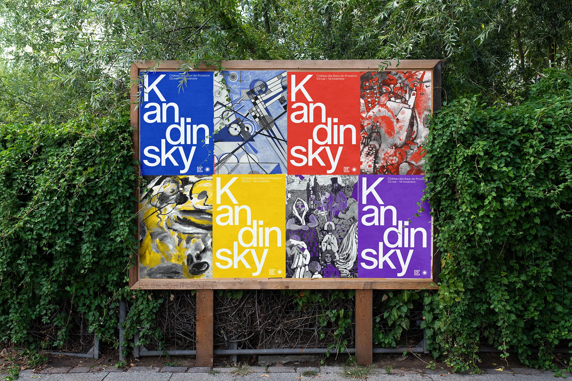 Outdoor display features six Kandinsky art posters in blue, black and white, yellow, red, and purple, surrounded by green foliage.