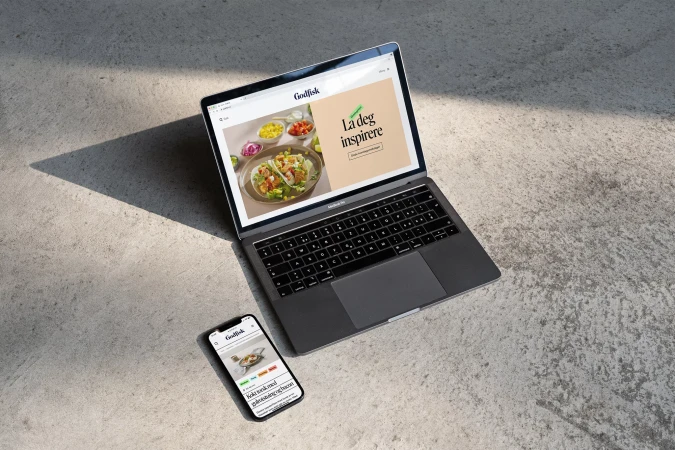 Laptop and smartphone displaying a Godlfisk website, with a dish image, on a grey surface with natural light and shadows.