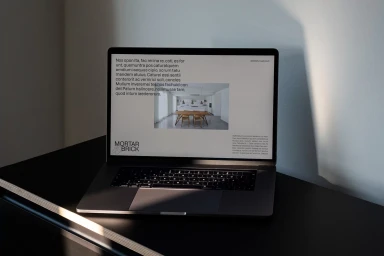 A dark laptop displaying Mortar&Brick branding on a black table with a wall casting sharp shadows.