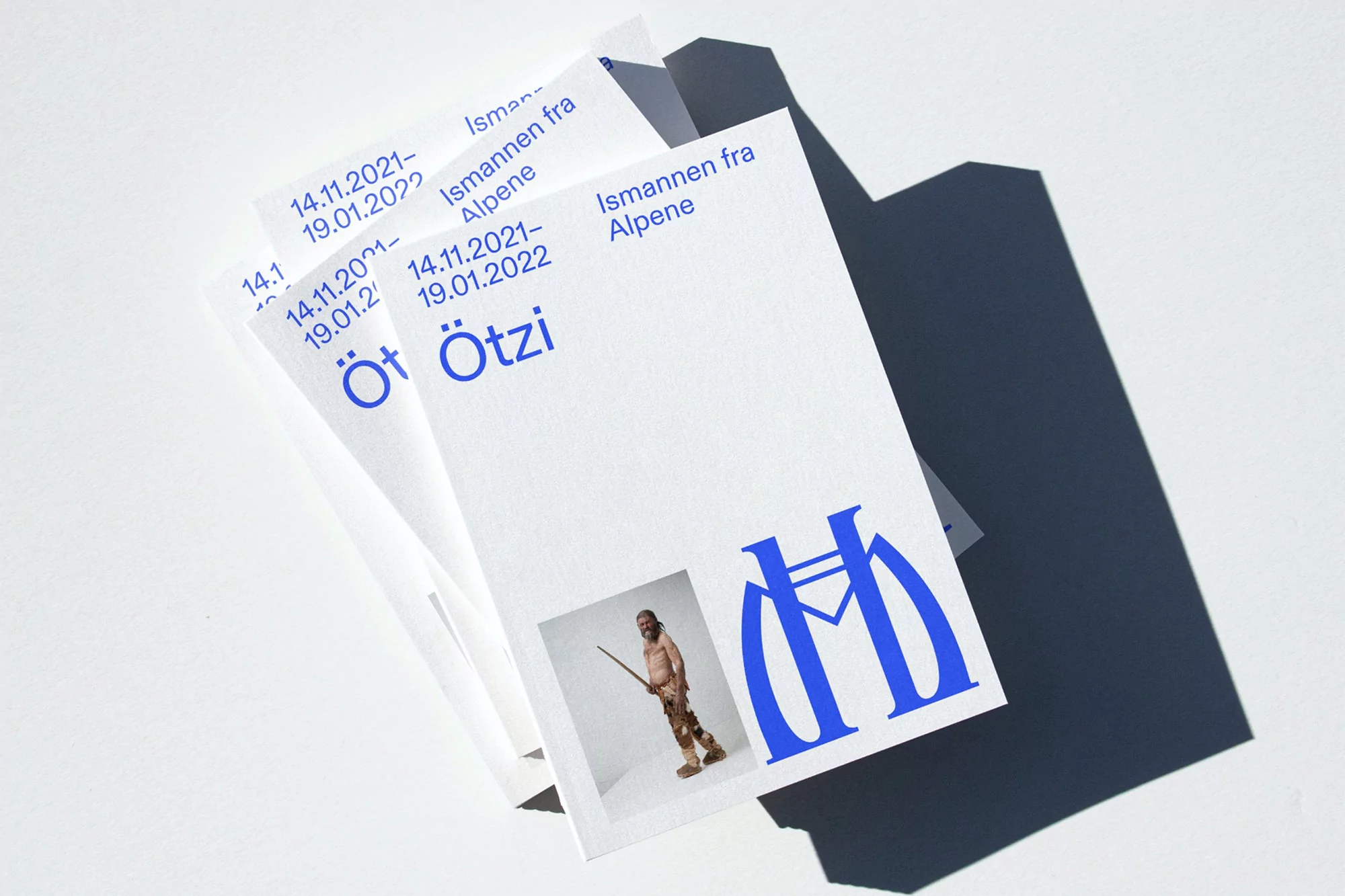 Flyers stacked in a pile show an image of the Iceman Ötzi, blue text, and museum logos against a white background with a shadow.