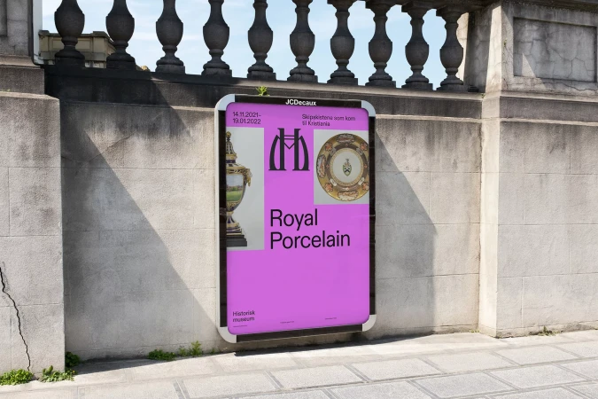 Outdoor advertising billboard on a city street displays a Royal Porcelain exhibit poster in bright purple and white, with stone wall backdrop.