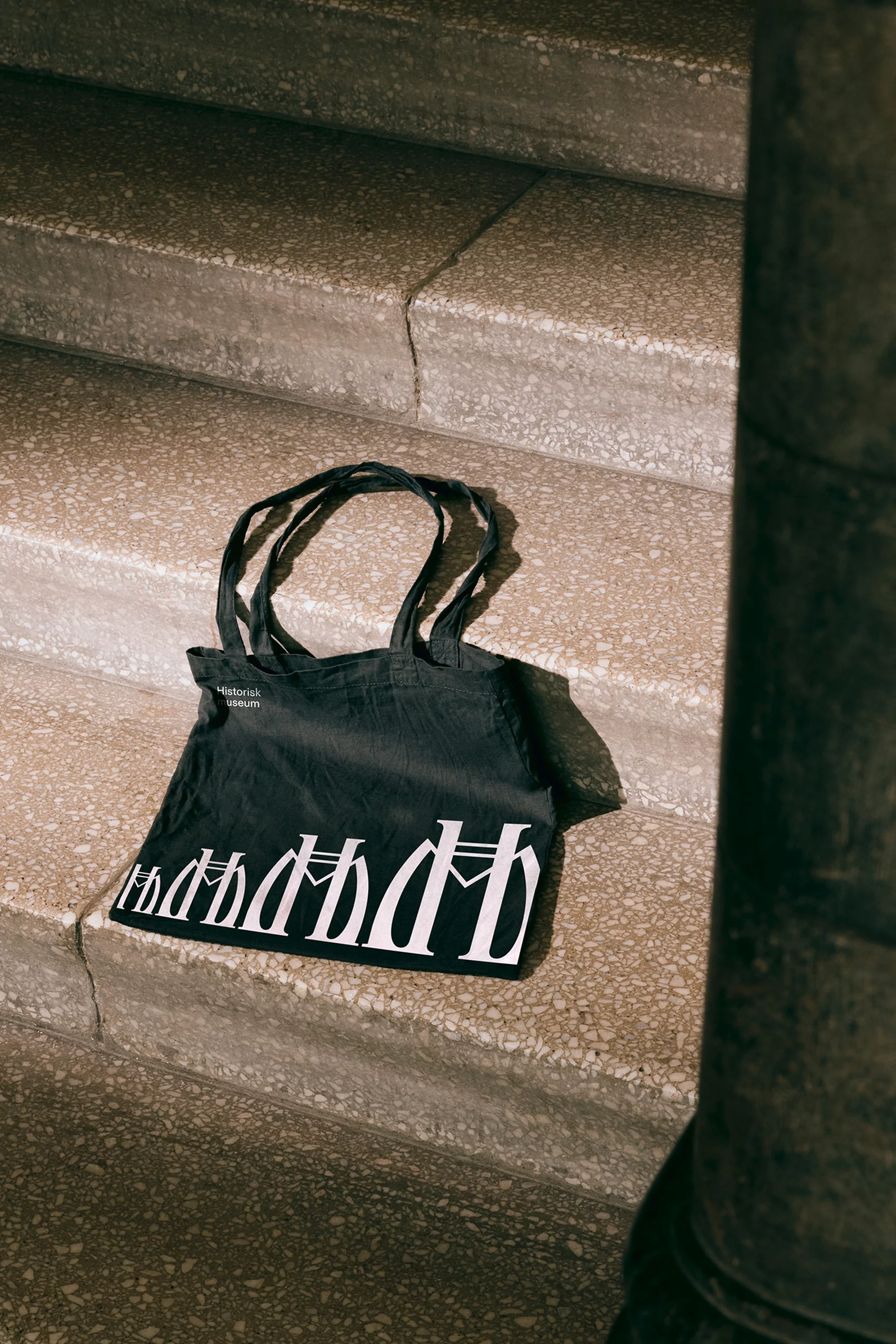 Black tote bag with white text on concrete stairs, bathed in natural light, casting a subtle shadow, showcasing minimalist design.