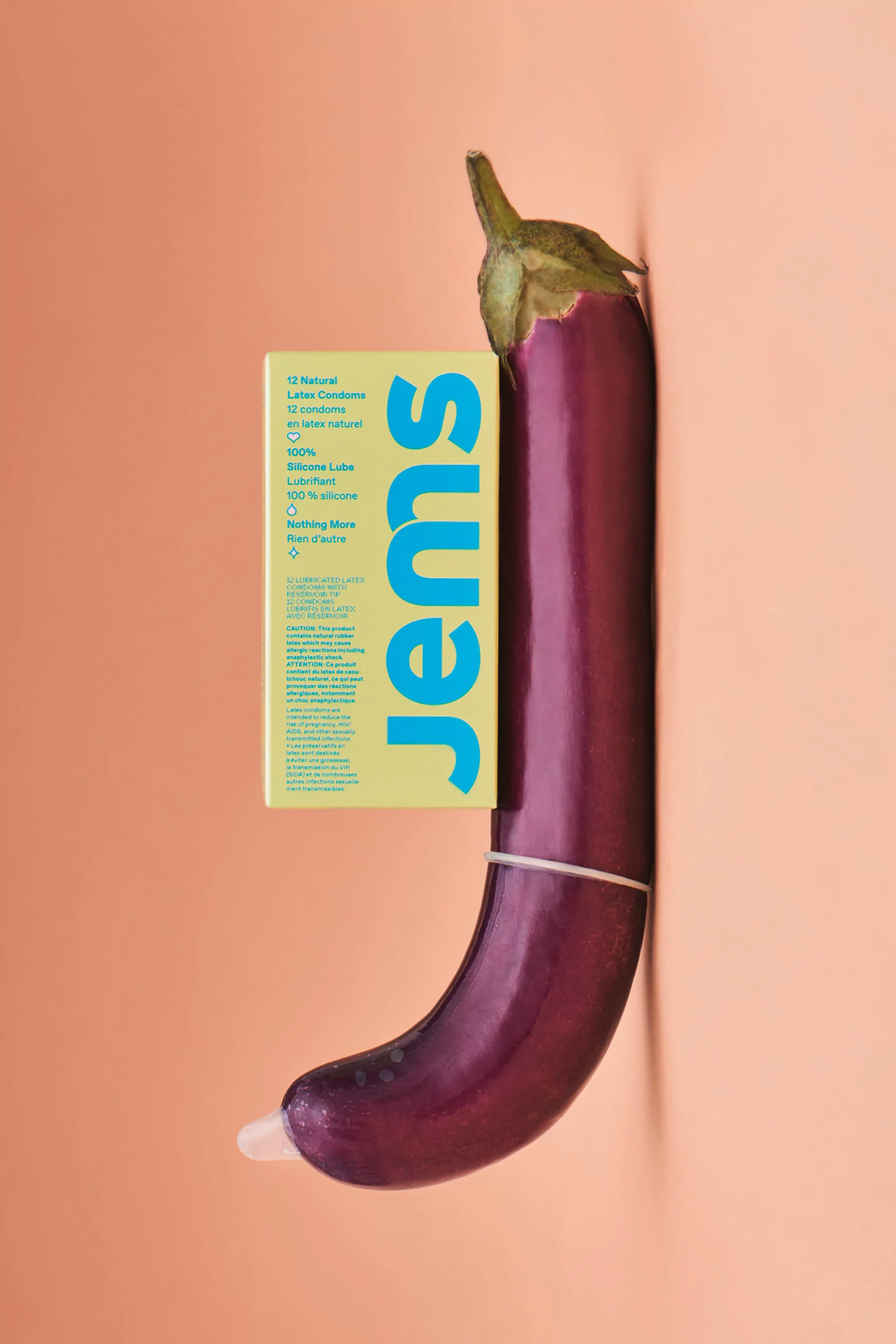 A still life features an eggplant with a condom, along with a box of Jems condoms, set against a warm peach-colored background.