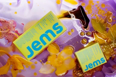 Bright yellow Jems product packaging boxes surrounded by yellow flowers and golden oil droplets on a light purple fabric background.