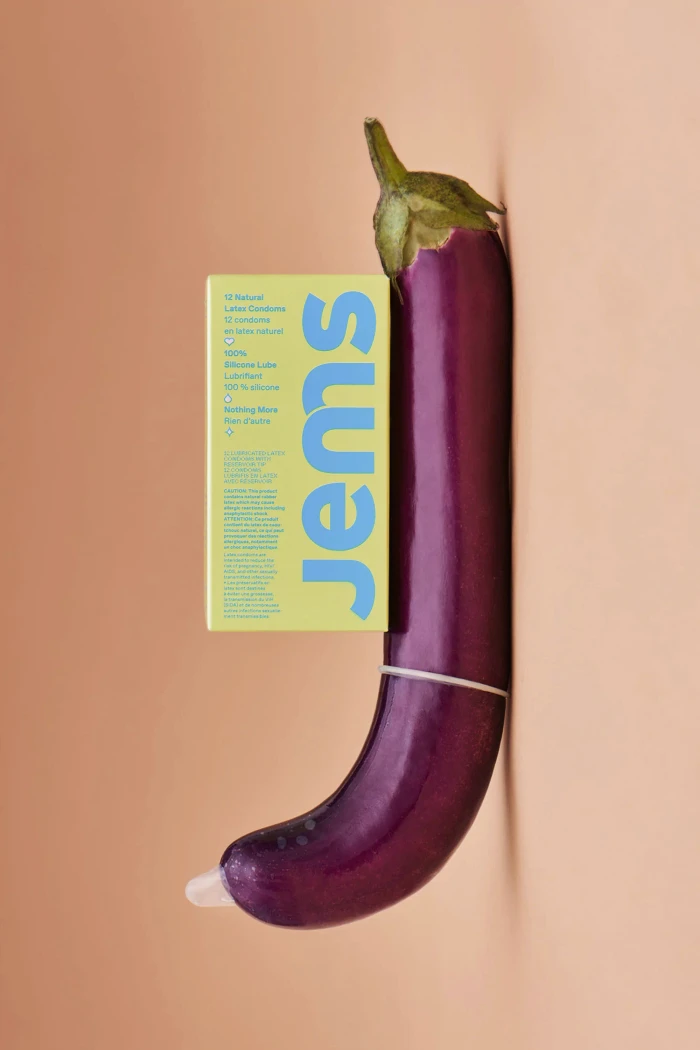 A purple eggplant with a condom on its tip and a yellow product box for Jems placed vertically against a peach background.