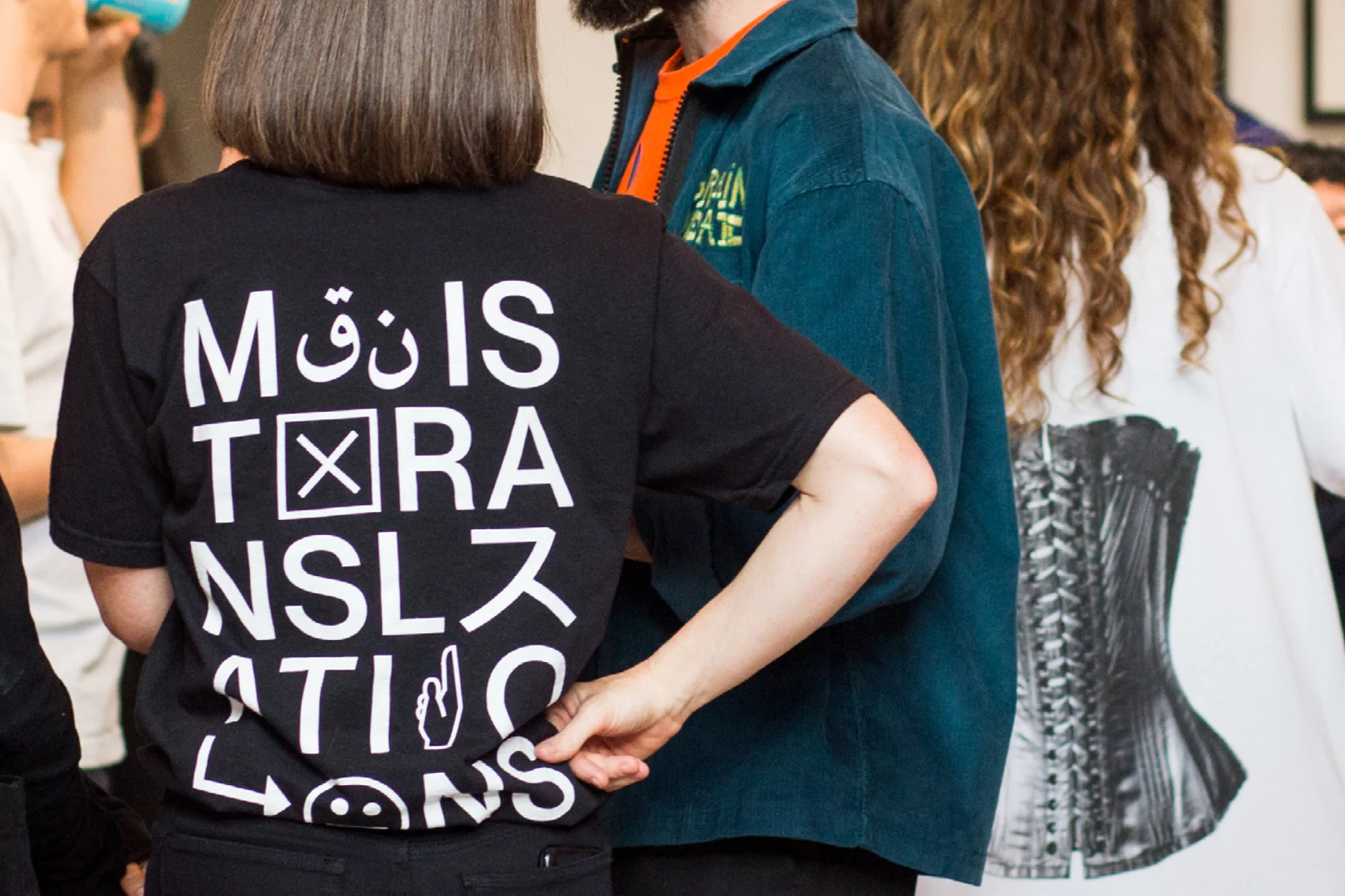 Back view of person wearing black t-shirt with white graphic text in various languages, standing in a group of people.