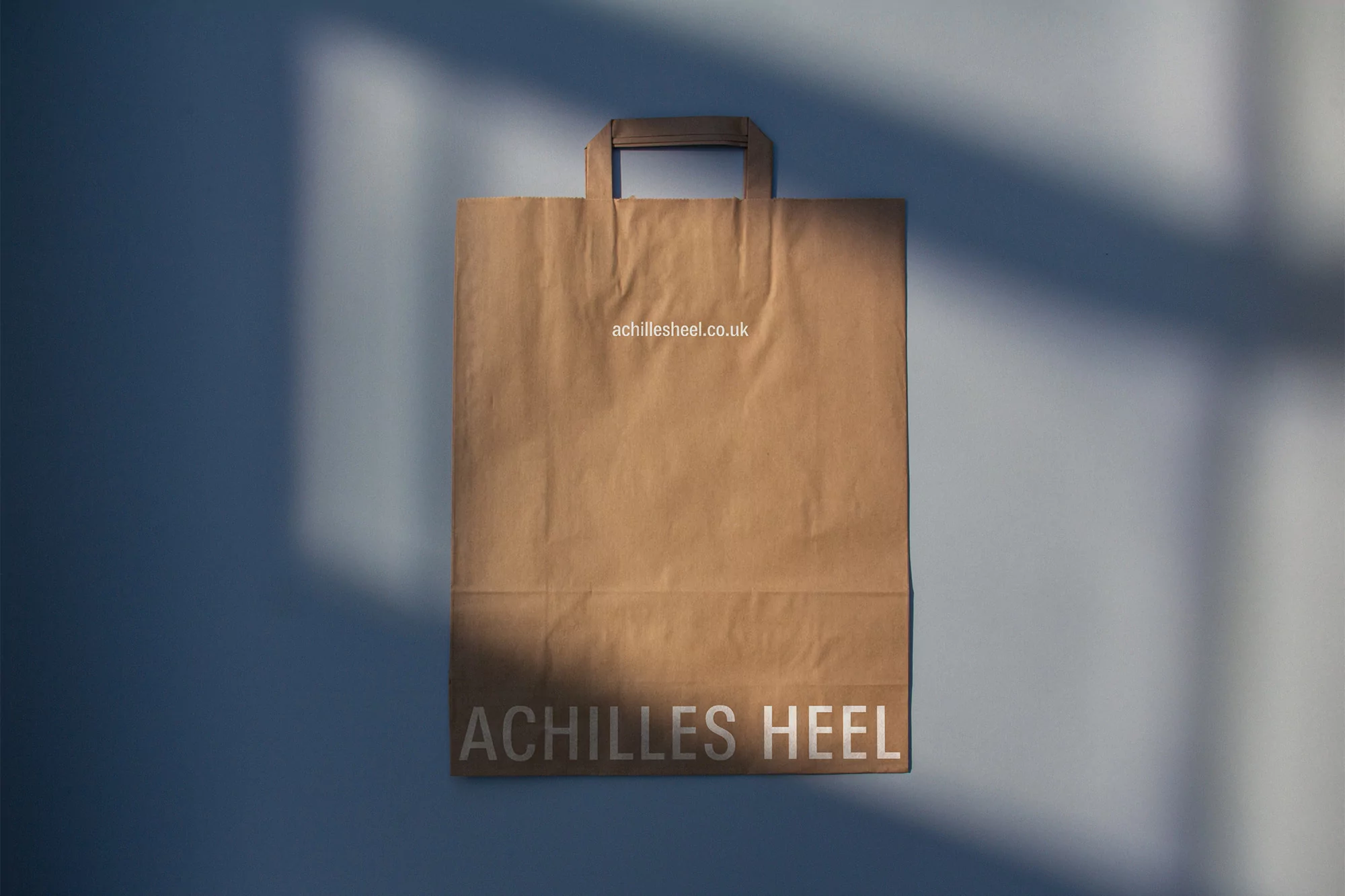 Kraft paper bag with handles, featuring 'achillesheel.co.uk' and 'ACHILLES HEEL' text, casting a shadow on a blue wall.