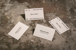 Four white business cards featuring Achilles Heel branding resting on a textured concrete surface.