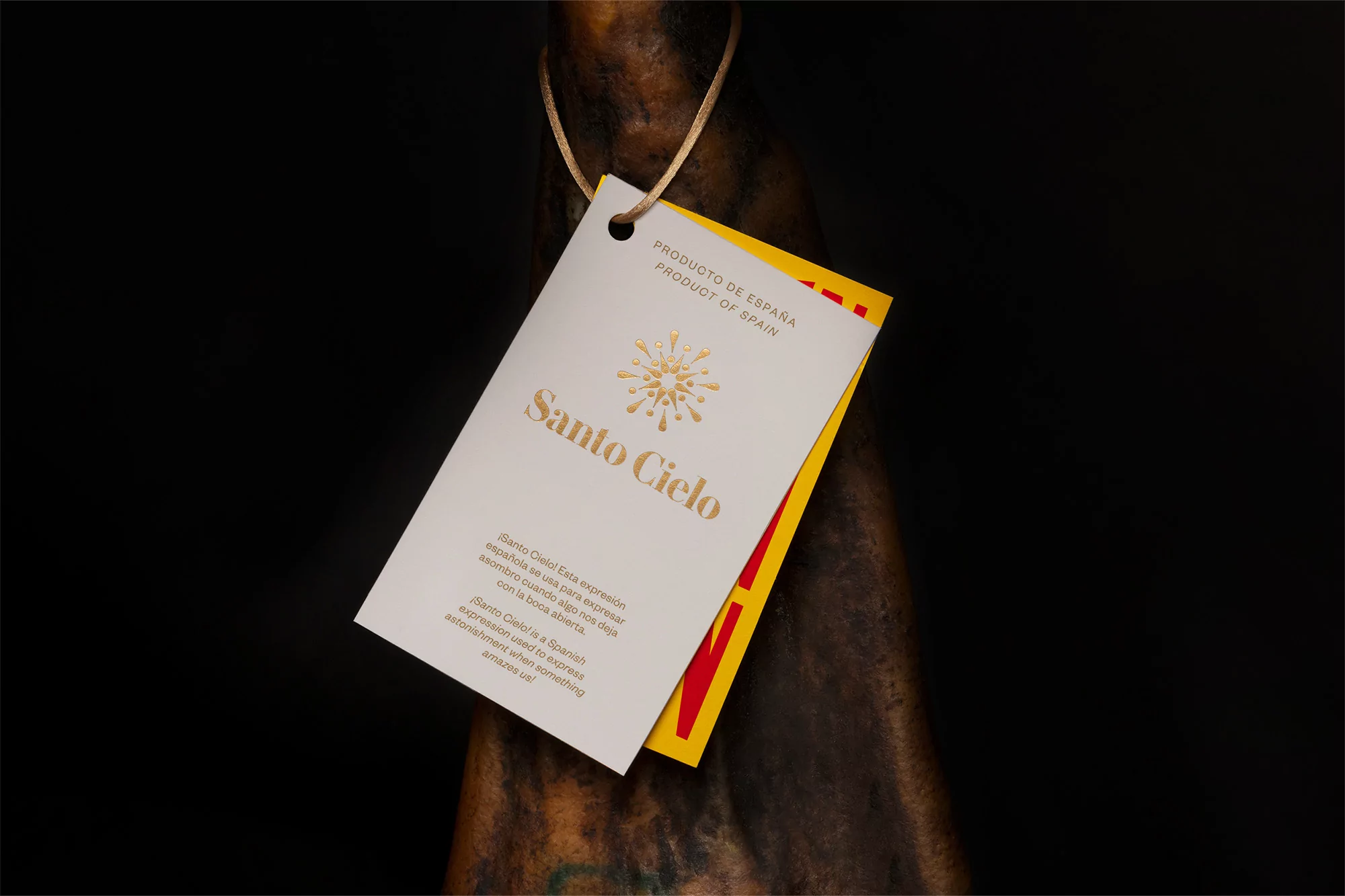 Close-up of a 'Santo Cielo' brand product tag in gold lettering, attached to a Spanish ham, against a dark background.