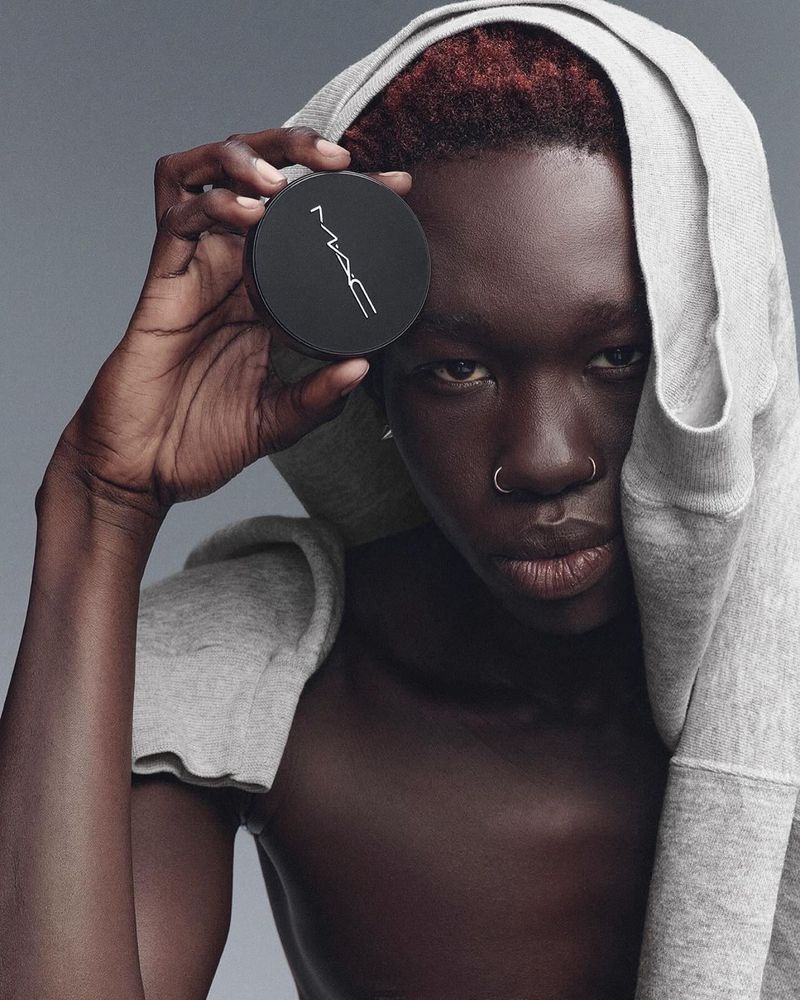 Dark-skinned young adult with nose rings posing in a grey hooded sweatshirt, holding a black MAC powder compact.