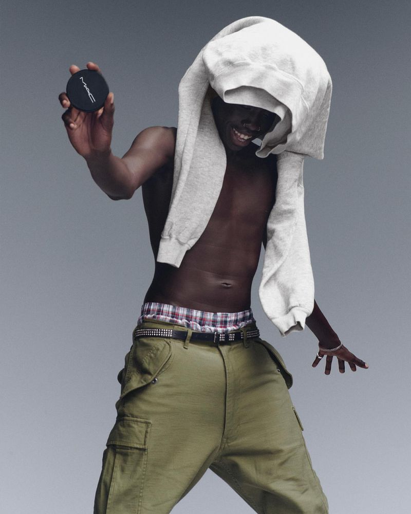 A black male model displays a MAC makeup compact, wearing baggy pants and a gray sweatshirt draped over his head in a studio shot.