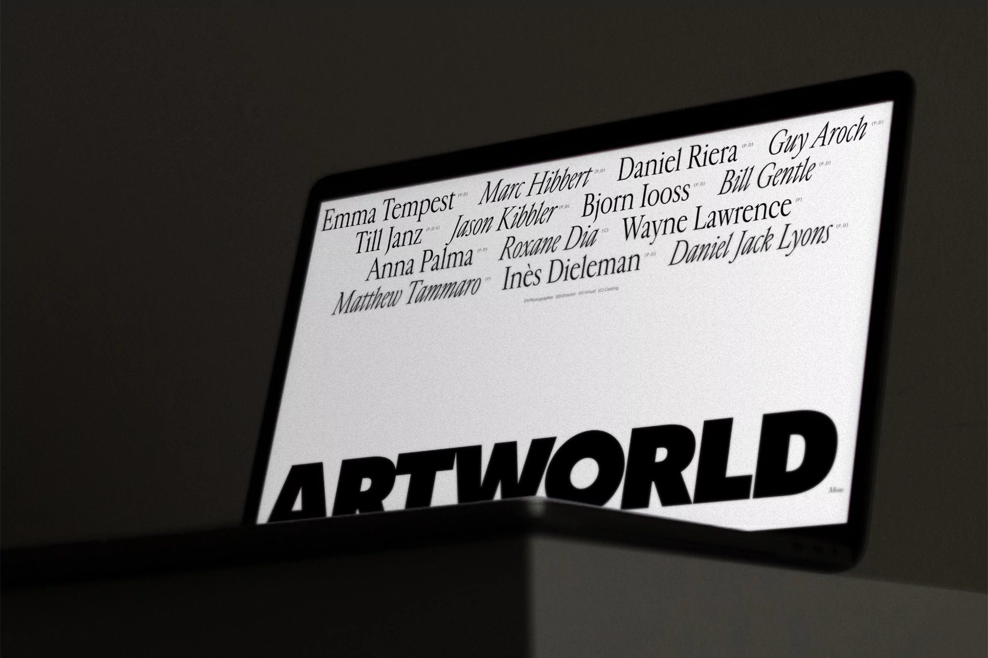 Laptop screen displaying 'ARTWORLD' and a list of names in a dark, indoor setting. The display is the focal point, with minimalist aesthetics.