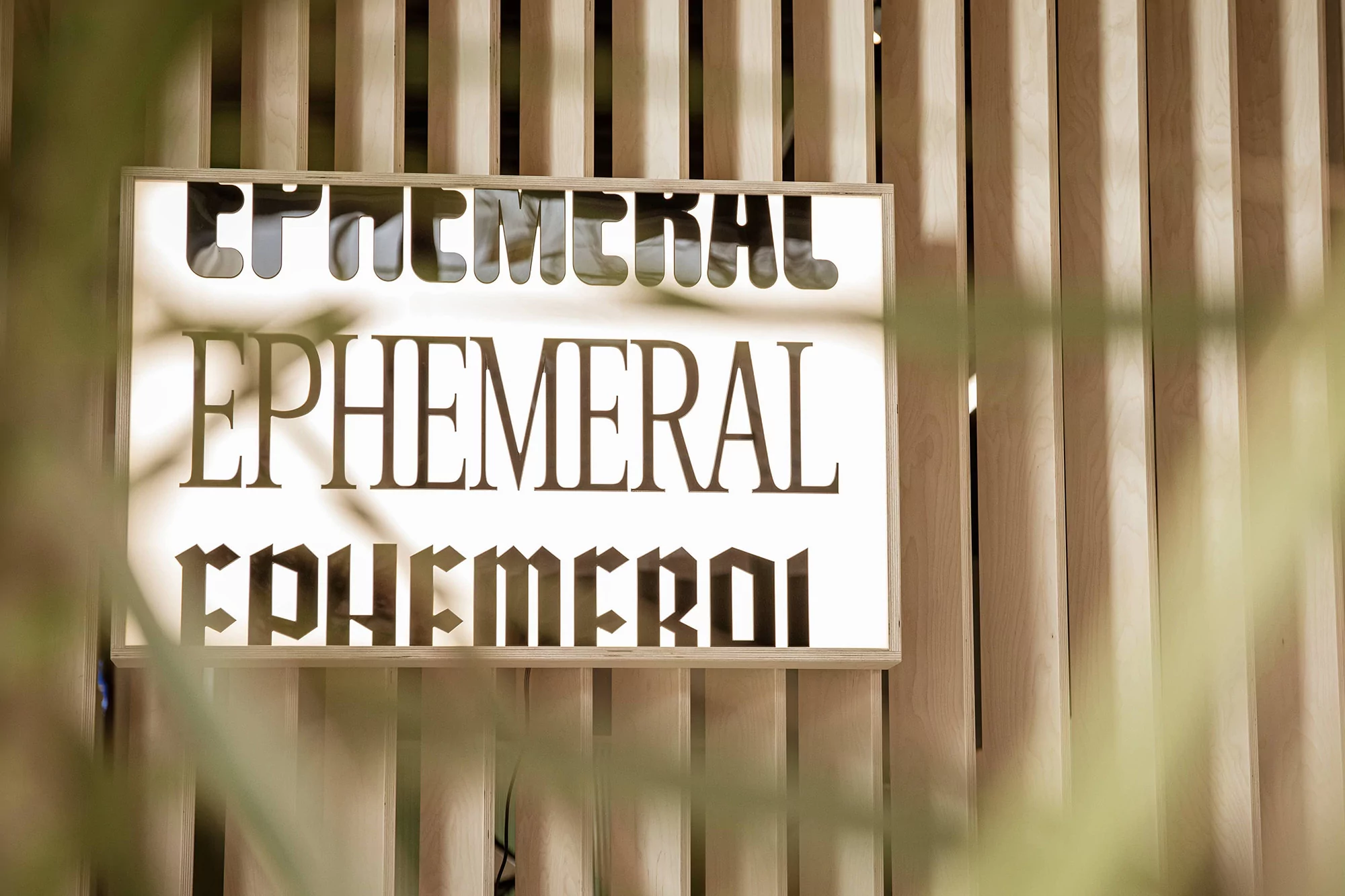 Close-up of a sign with the word 'Ephemeral' in a decorative font, mounted on a wooden slat wall, with green foliage in the foreground.