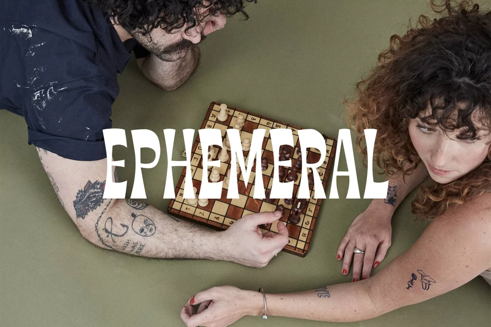 A man and woman are playing chess, captured from an overhead view, showcasing their arms, tattoos, and the chess board.
