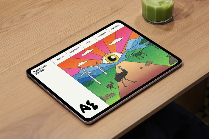 Tablet displays colorful, illustrative website showing animals and wind turbines, resting on a wood desk next to a green drink.