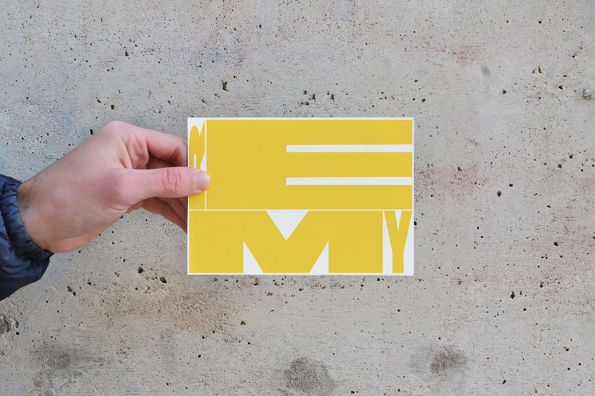 Hand holds a yellow and white graphic card against a textured gray wall, featuring bold typography and a minimalist design.