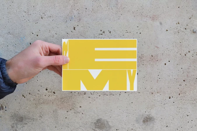 Hand holding a white card featuring yellow graphic typography against a textured concrete wall.