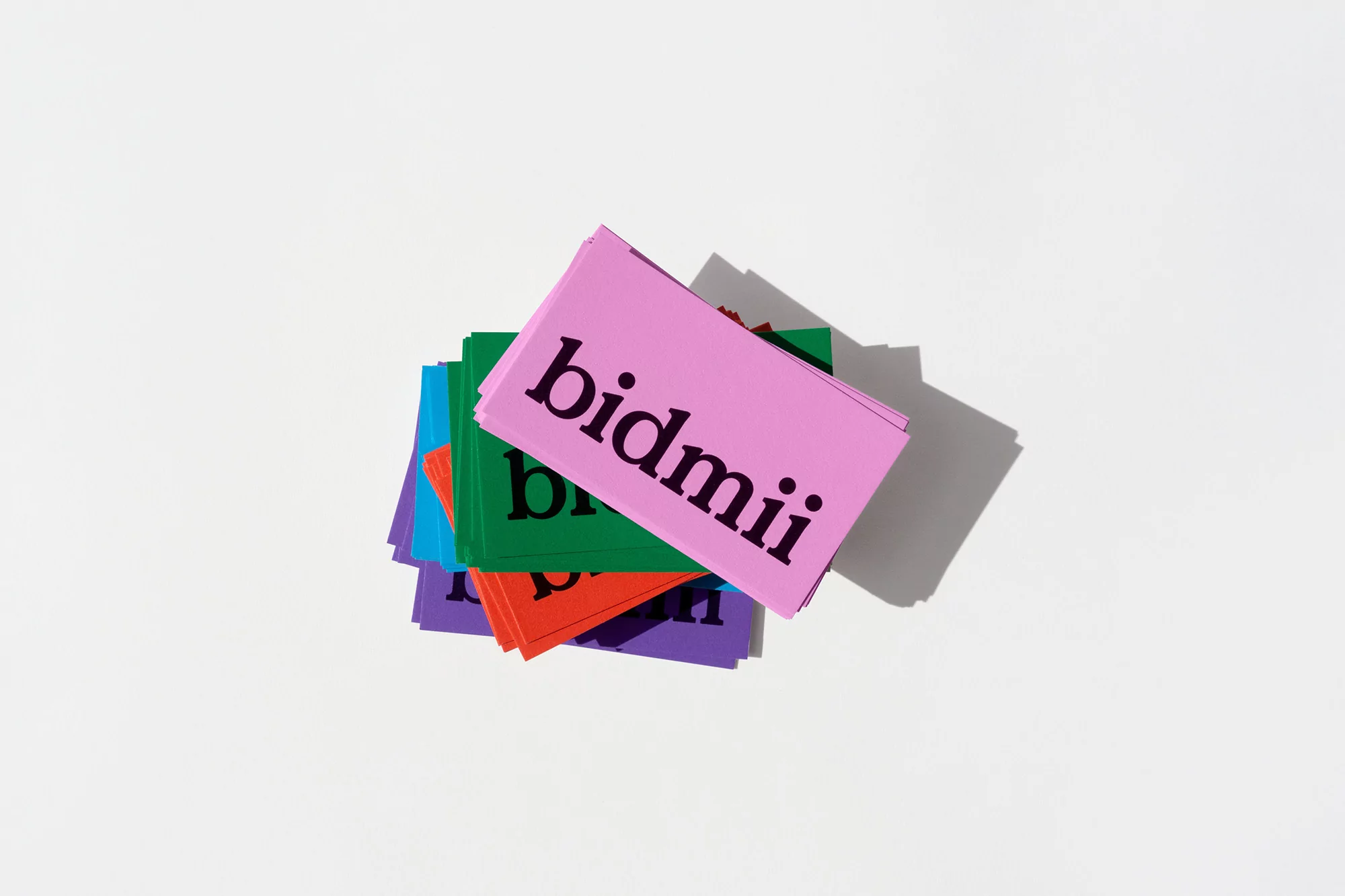 Overhead shot of a stack of colorful cards, each displaying the word 'bidmii' in bold, black letters, set against a bright white backdrop.