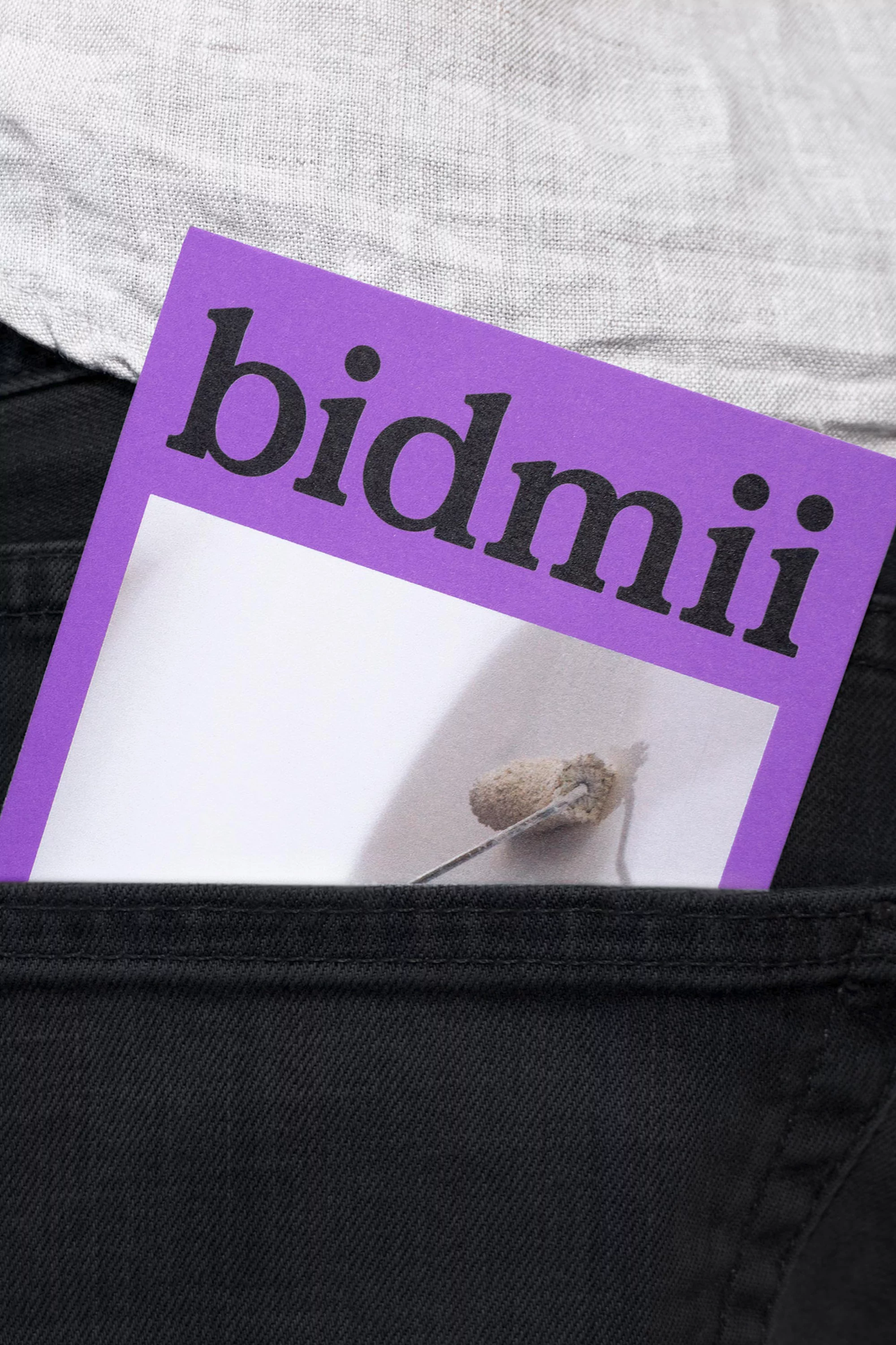 Close-up shot of a purple card with the text "bidmii" sticking out of a black pants pocket, set against a light grey shirt.