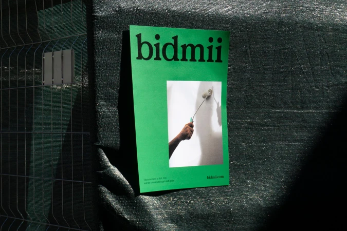 Promotional image featuring a green bidmii flyer attached to a chain-link fence, showcasing a hand painting a wall with a roller.