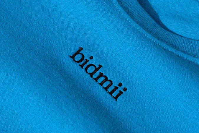 Close-up of a turquoise T-shirt with the black embroidered text 'bidmii' on it, in a studio setting with diffused lighting.