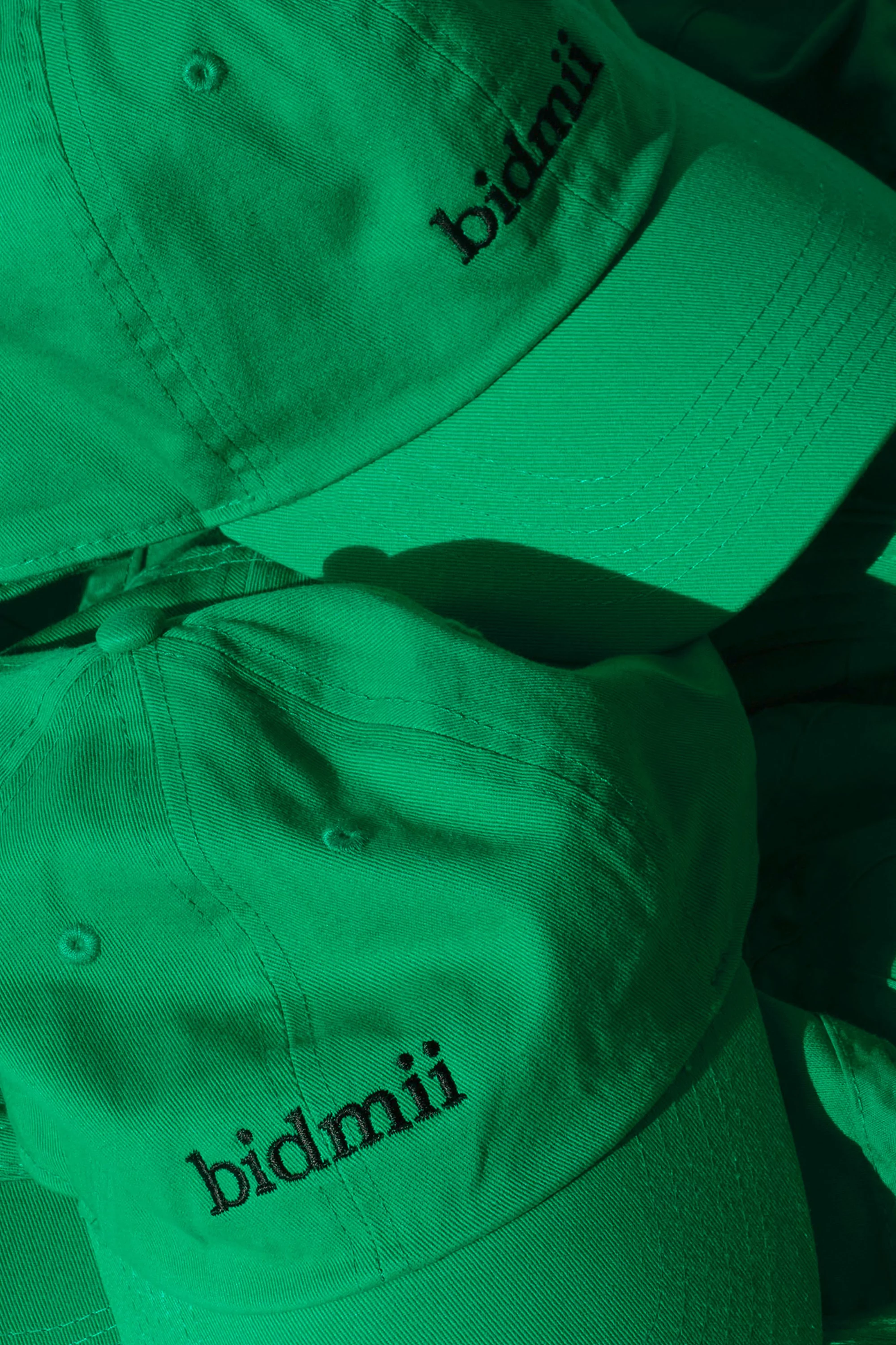 Pile of light green baseball caps with black embroidered logo. Hats are stacked with shadows. The hats are the dominant subject.