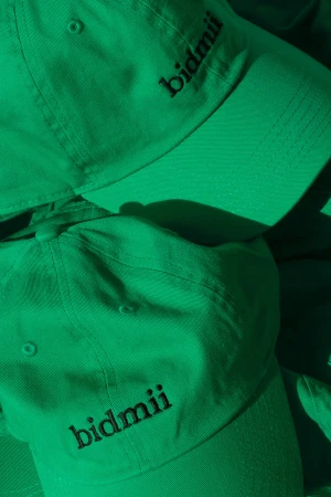 Pile of light green baseball caps with black embroidered logo. Hats are stacked with shadows. The hats are the dominant subject.