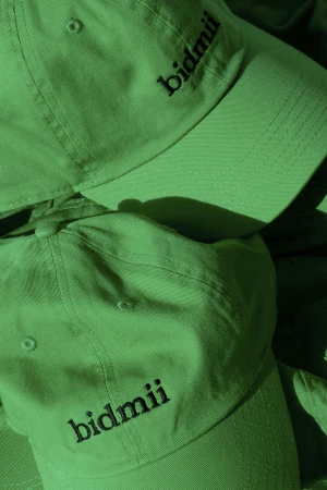 Green baseball caps stacked with embroidered black text on the front for Bidmii brand identity.