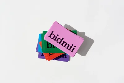 Stack of colourful printed cards featuring the Bidmii brand identity on white seamless background